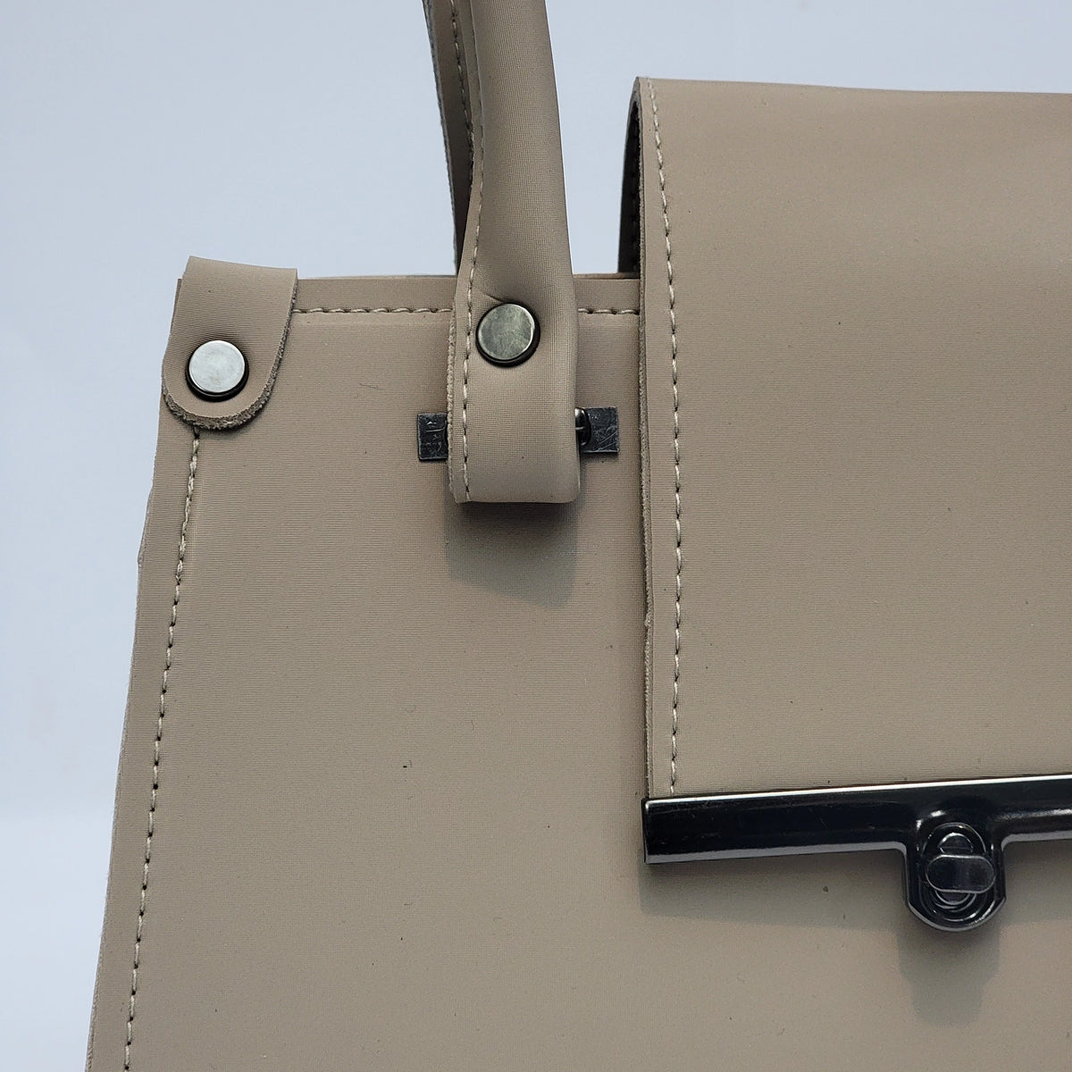 Close-up of a structured beige handbag with a top handle, visible stitching, metal rivets, and a black metal bar clasp on a light gray background.