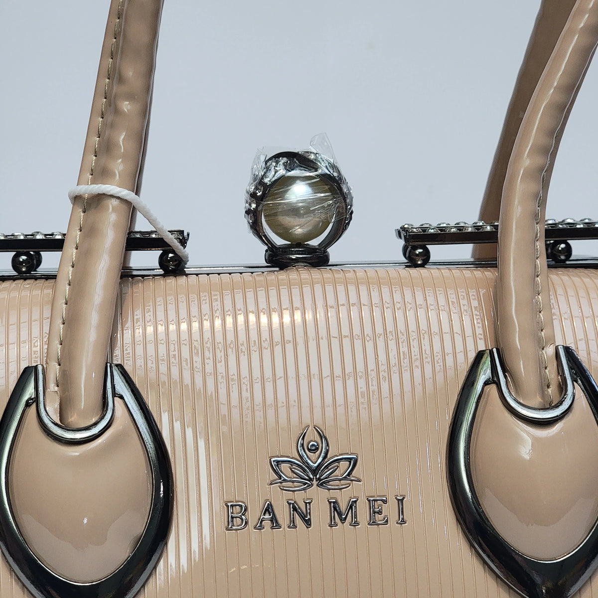 Close-up of a beige textured handbag with glossy taupe handles, silver-tone hardware, and the BAN MEI logo on the front, set against a light gray background.