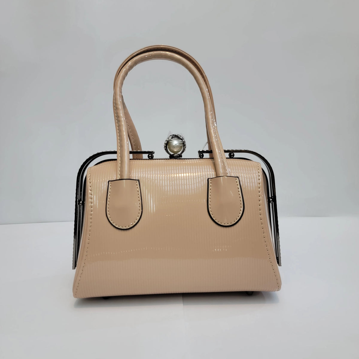 A structured beige patent leather handbag with double top handles, a black metal frame, and a pearl-embellished clasp, shown on a light gray background.