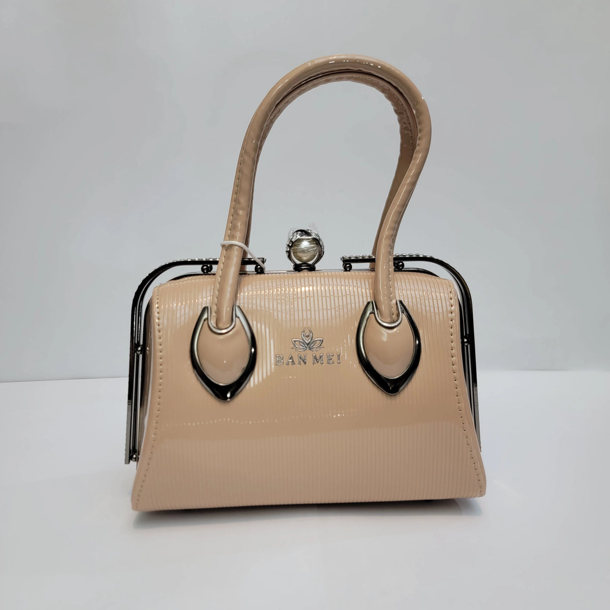 A structured handbag in beige patent leather with a glossy finish, featuring a double top handle, black metal frame, and a pearl clasp closure. The brand name 'JEAN ME!' is printed on the front.