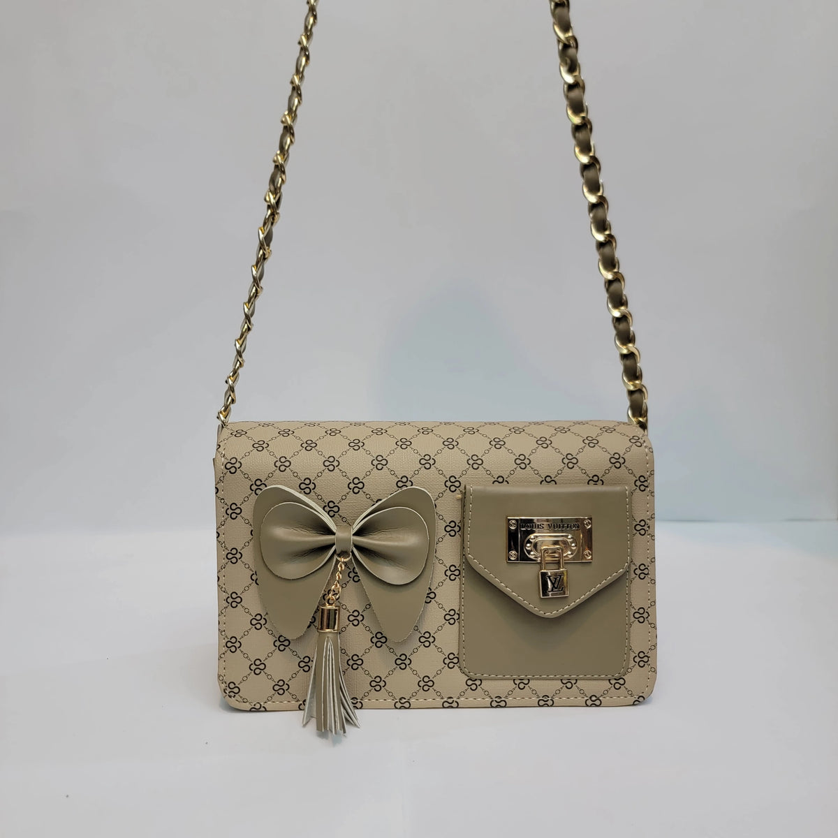A beige shoulder bag with a repeating monogram pattern, featuring a large matching fabric bow with a tassel on the front pocket, a gold-tone clasp, and a gold chain strap.