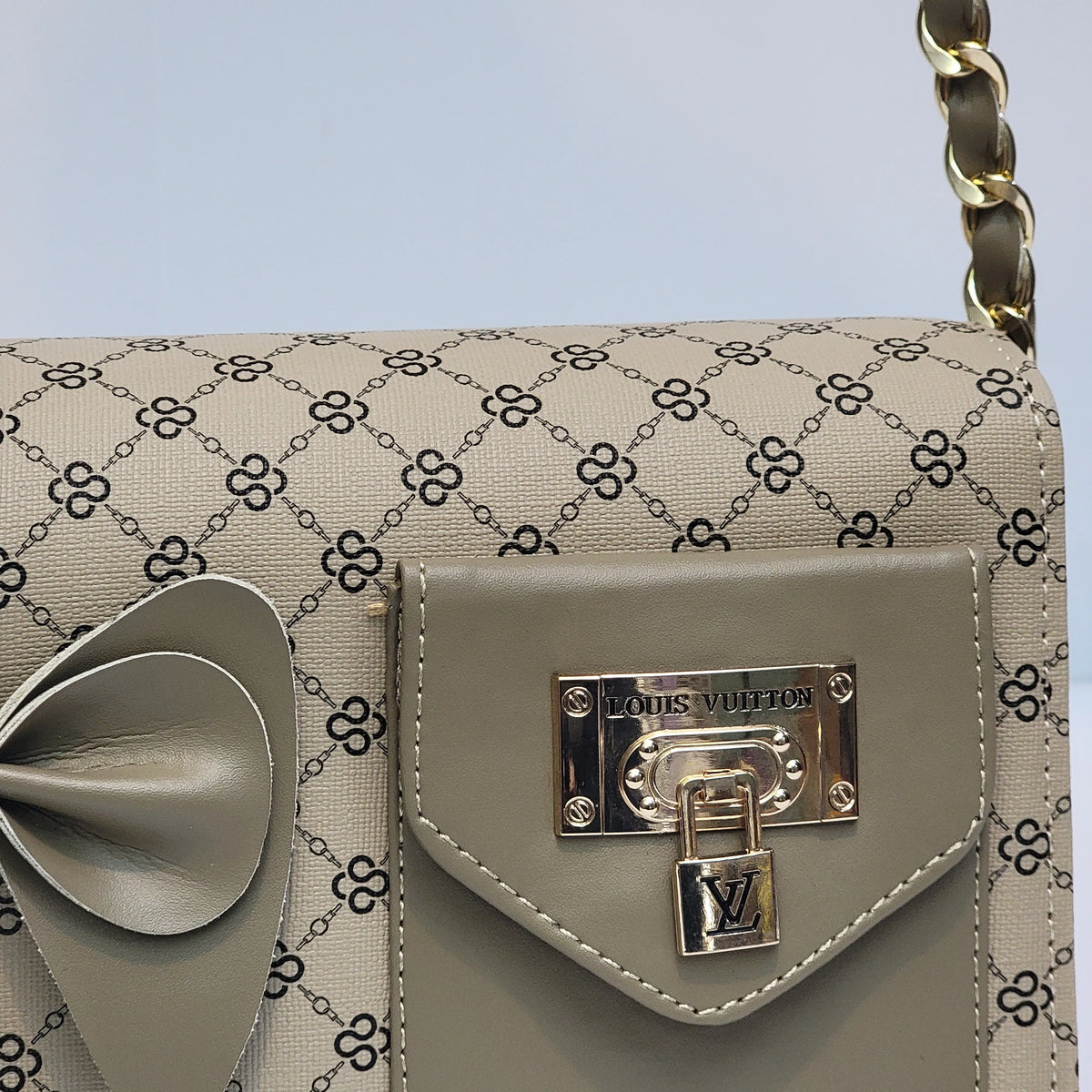 Close-up of a Louis Vuitton handbag featuring beige and black GG Supreme canvas, a taupe leather front flap with a gold-tone Louis Vuitton padlock clasp, and a gold chain strap against a light blue-gray background.