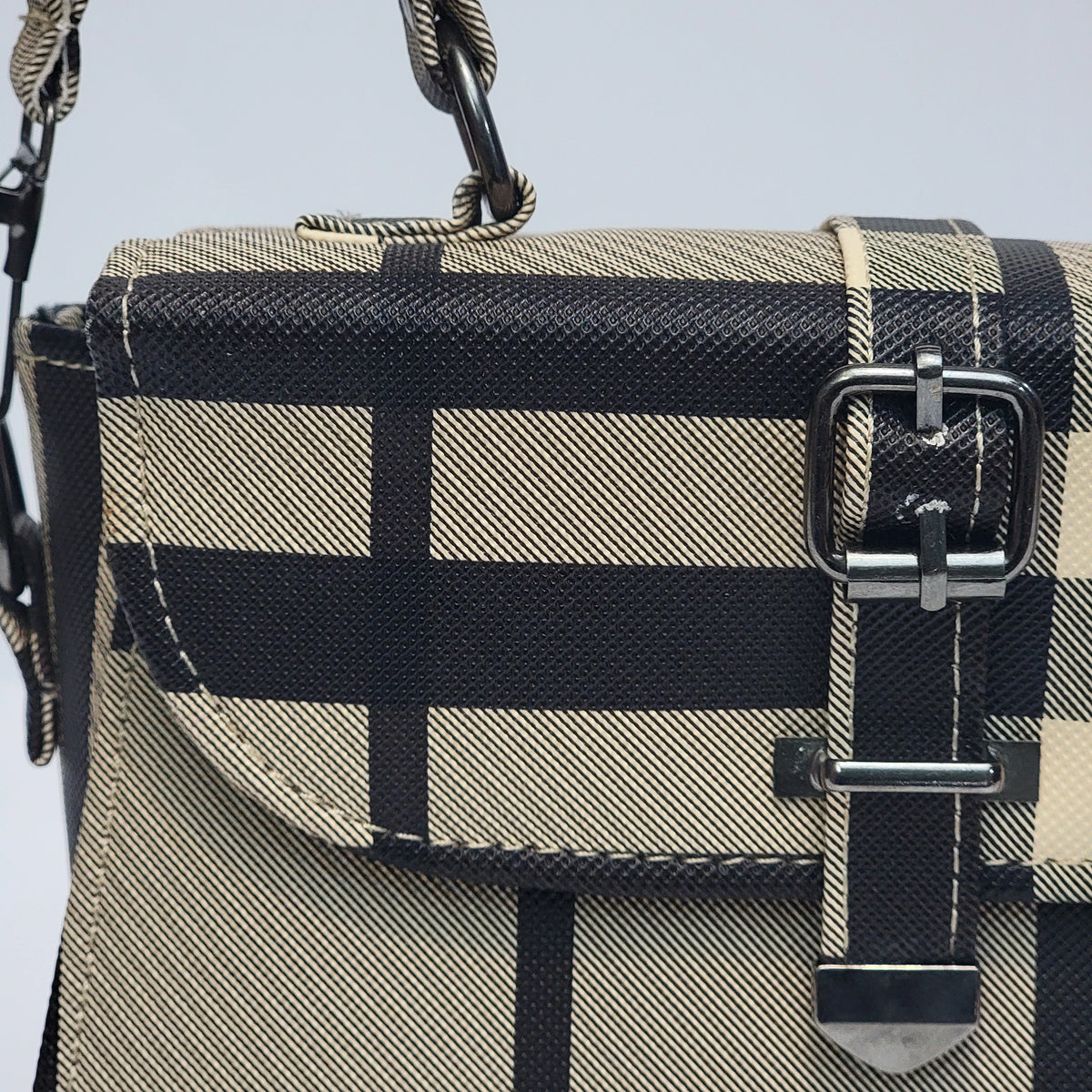 Close-up of a structured handbag with a beige and black plaid pattern, featuring a flap front, silver-tone buckle closure, and rope-style strap details.