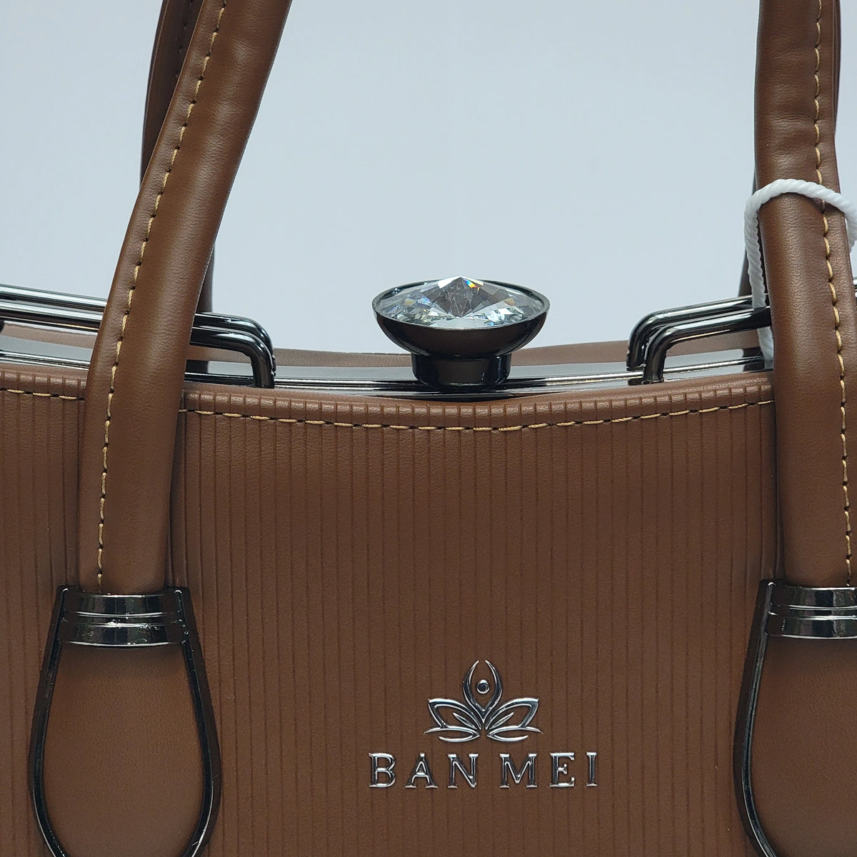 Close-up of a brown textured leather handbag with gold-toned metal hardware, a clear faceted top gem, and the BAN MEI logo embossed on the front.