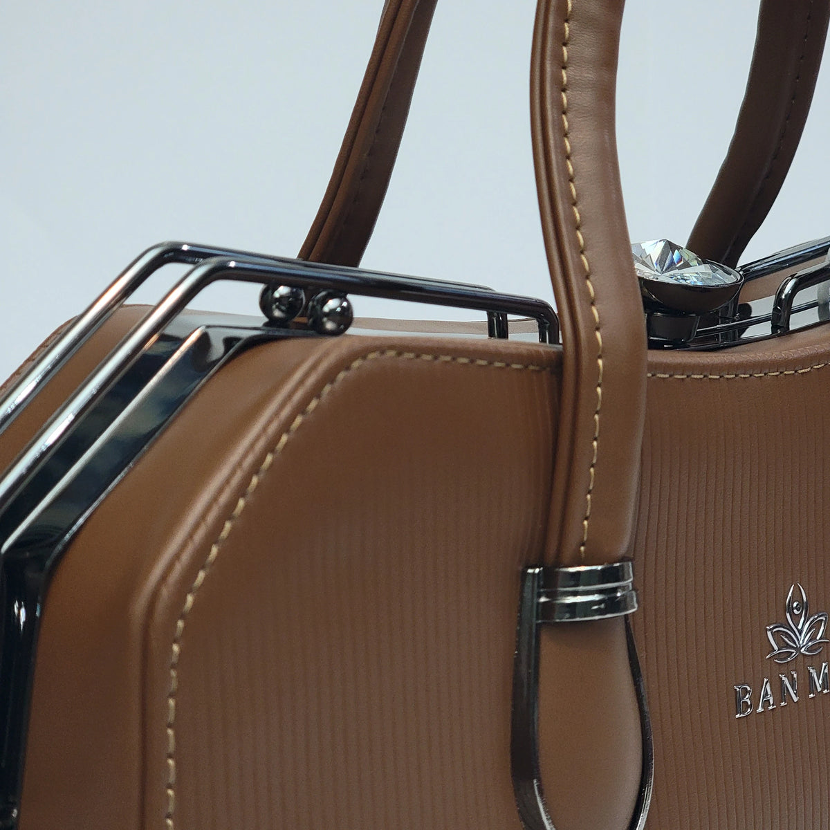 Close-up of a brown textured leather handbag with cream stitching, dark metal frame details, and a silver-tone logo on the lower right.