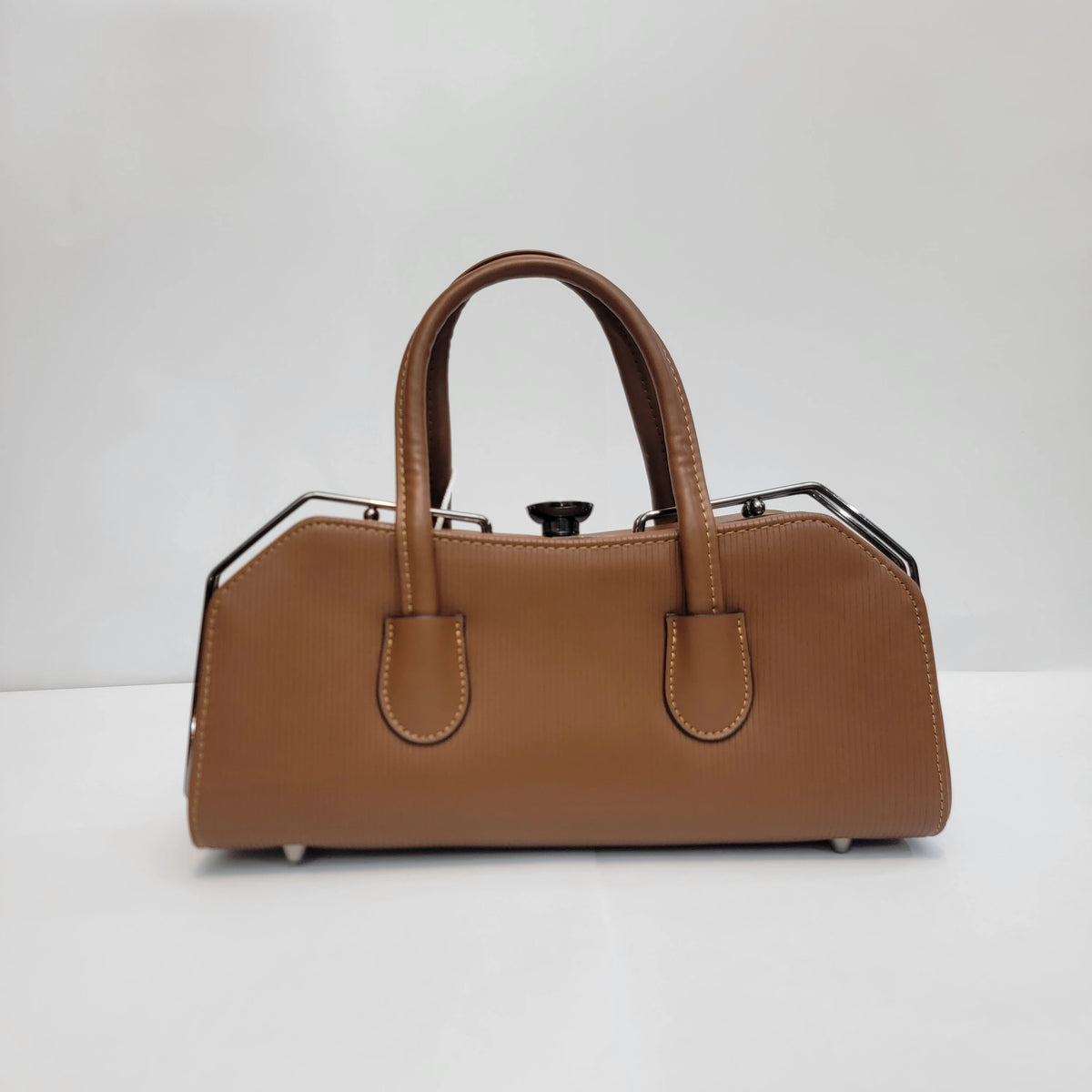 A structured tan leather handbag with double top handles, a top clasp closure, and black metal side accents, photographed against a plain light gray background.
