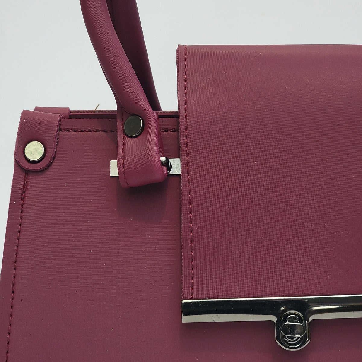 Close-up of a burgundy leather handbag showing the flap closure, top handle attachment, and silver-tone hardware against a light gray background.