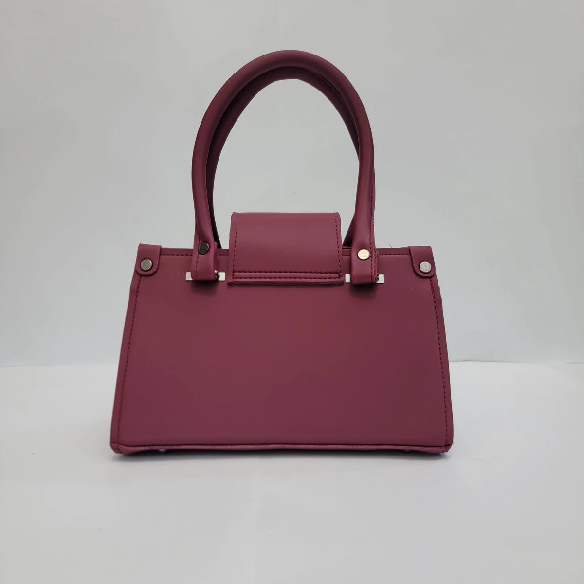 A structured burgundy handbag with a top handle, silver-tone hardware, and a front flap, photographed against a neutral light gray background.