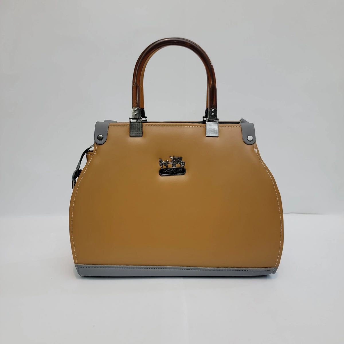 A structured Coach handbag in tan leather with brown handles, silver-tone hardware, and a black horse-and-carriage logo plaque on the front, photographed against a plain light gray studio background.
