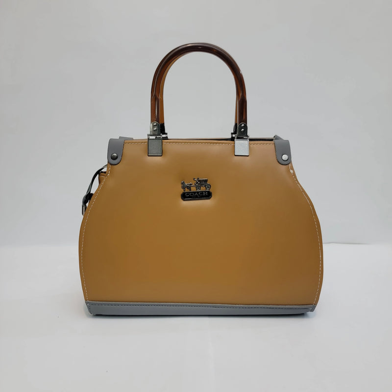 A structured Coach handbag in tan leather with brown handles, silver-tone hardware, and a black horse-and-carriage logo plaque on the front, photographed against a plain light gray studio background.