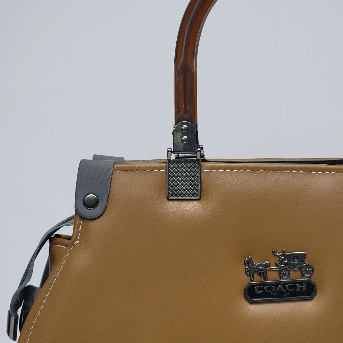 Close-up of a Coach handbag in tan leather with a dark brown wooden top handle, gray leather strap detail, and a black metal Coach logo plaque on the front.