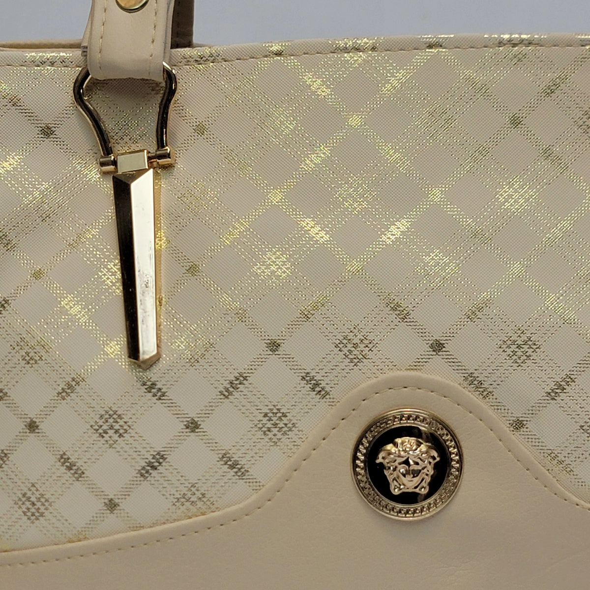 Close-up of a Versace handbag in off-white faux leather with a gold-toned diamond pattern, featuring a gold-tone Medusa emblem and a decorative strap clasp.