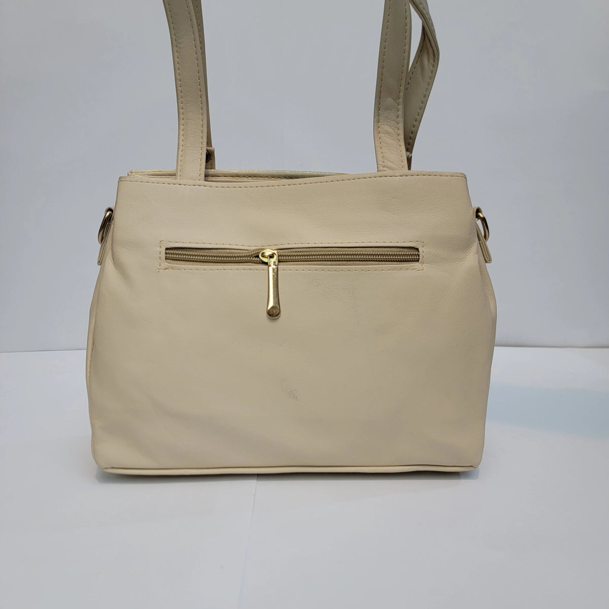 A beige faux-leather handbag with a front zippered pocket and gold-tone hardware, shown against a plain white studio background.