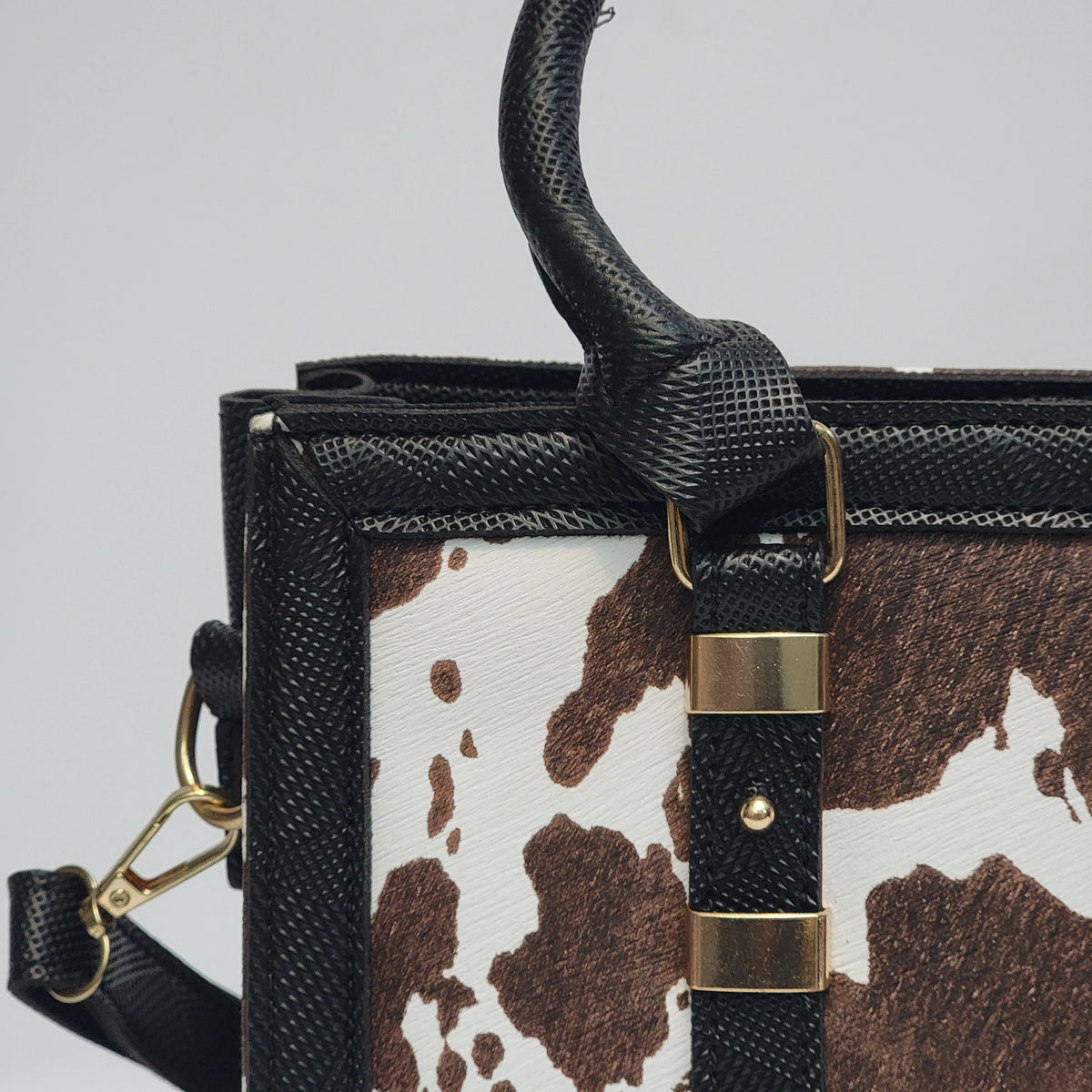 Close-up of a structured handbag featuring a brown and white cow print front panel, black textured leather trim and handle, and gold-tone hardware.