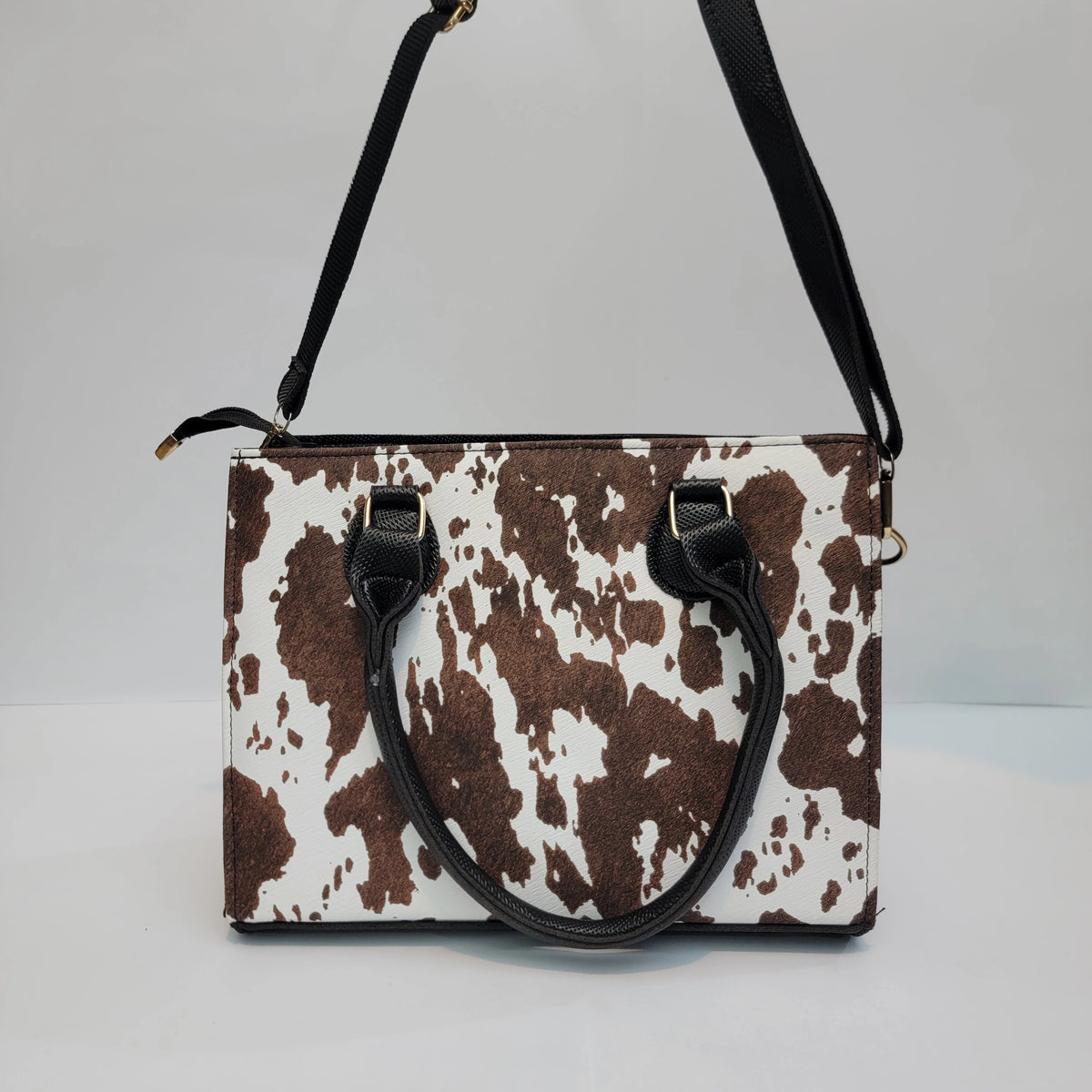 A cow-print handbag with brown and white spots, black leather handles and shoulder strap, and gold-tone hardware, photographed against a plain light gray background.