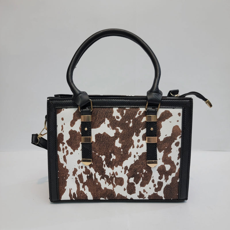 A structured handbag with a brown and white cow print front panel, black textured leather trim and top handle, and gold-tone hardware, photographed against a plain light gray background.