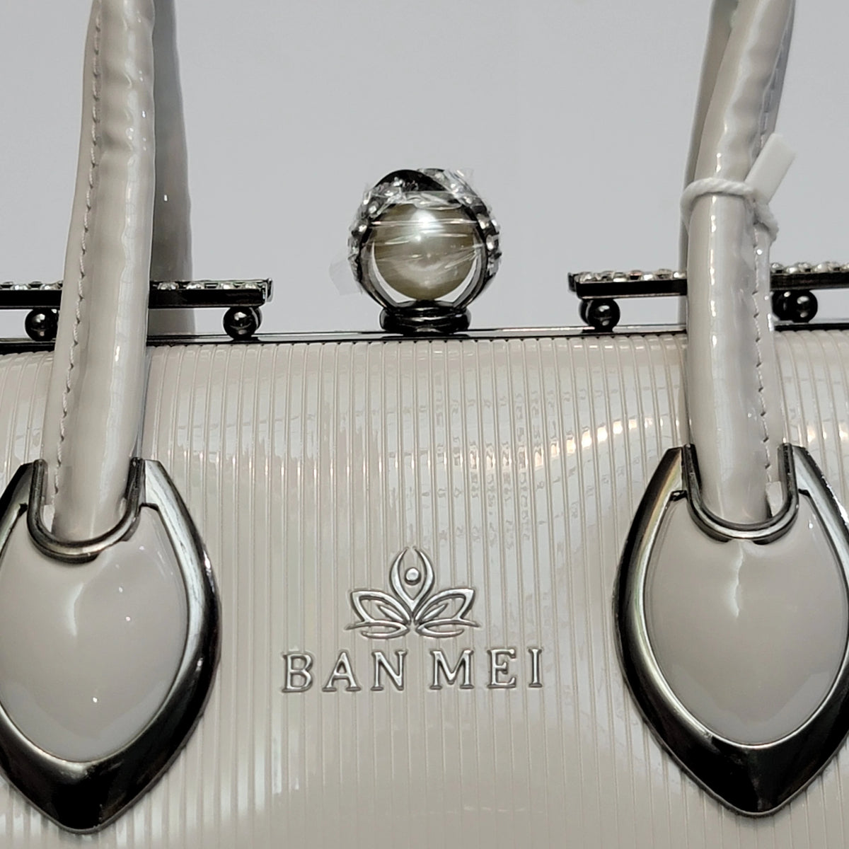 Close-up of a Ban Mei handbag in glossy off-white patent leather with silver-tone hardware, a central round pearl-like ornament, and the Ban Mei logo embossed on the front.
