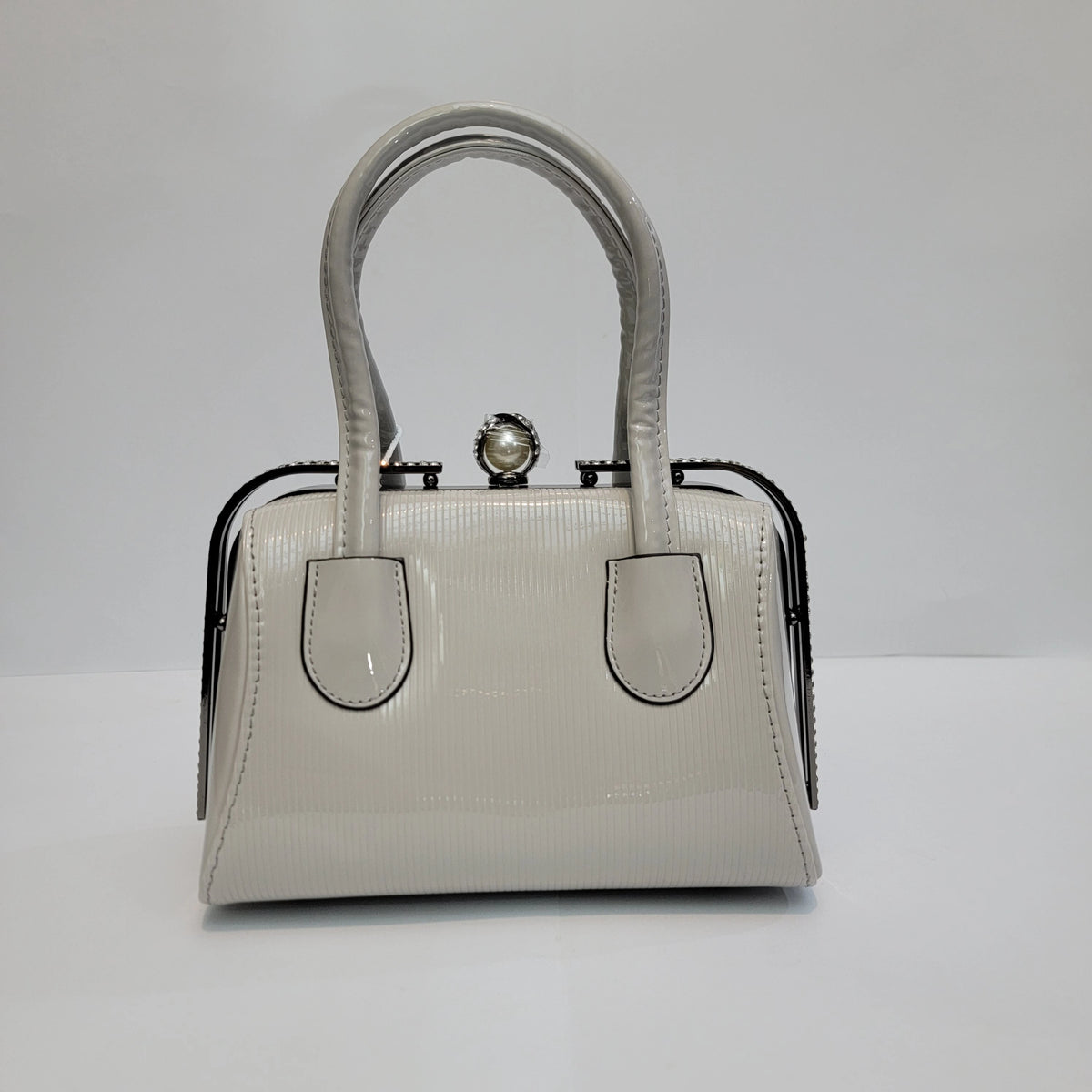 A small structured handbag in glossy light gray patent leather with double top handles, a kiss-lock clasp closure, and black metal side trim, photographed against a plain light gray studio background.