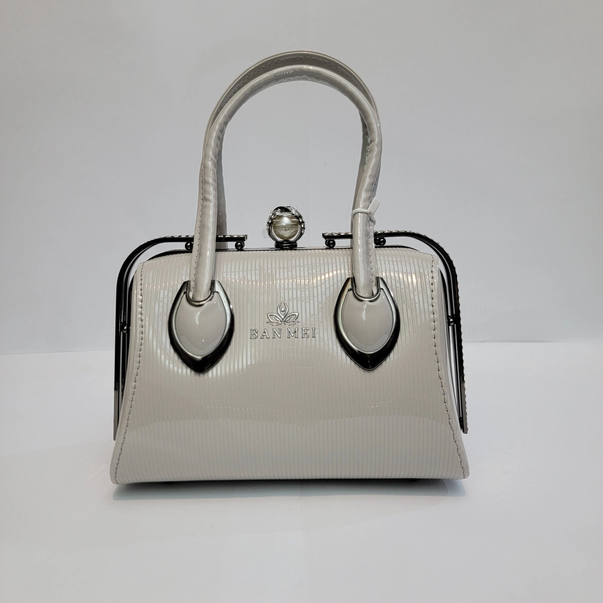 A small structured handbag in glossy white textured leather with black metal frame detailing, dual top handles, and a pearl-like clasp closure. The brand name 'BAJAMI' is printed on the front.