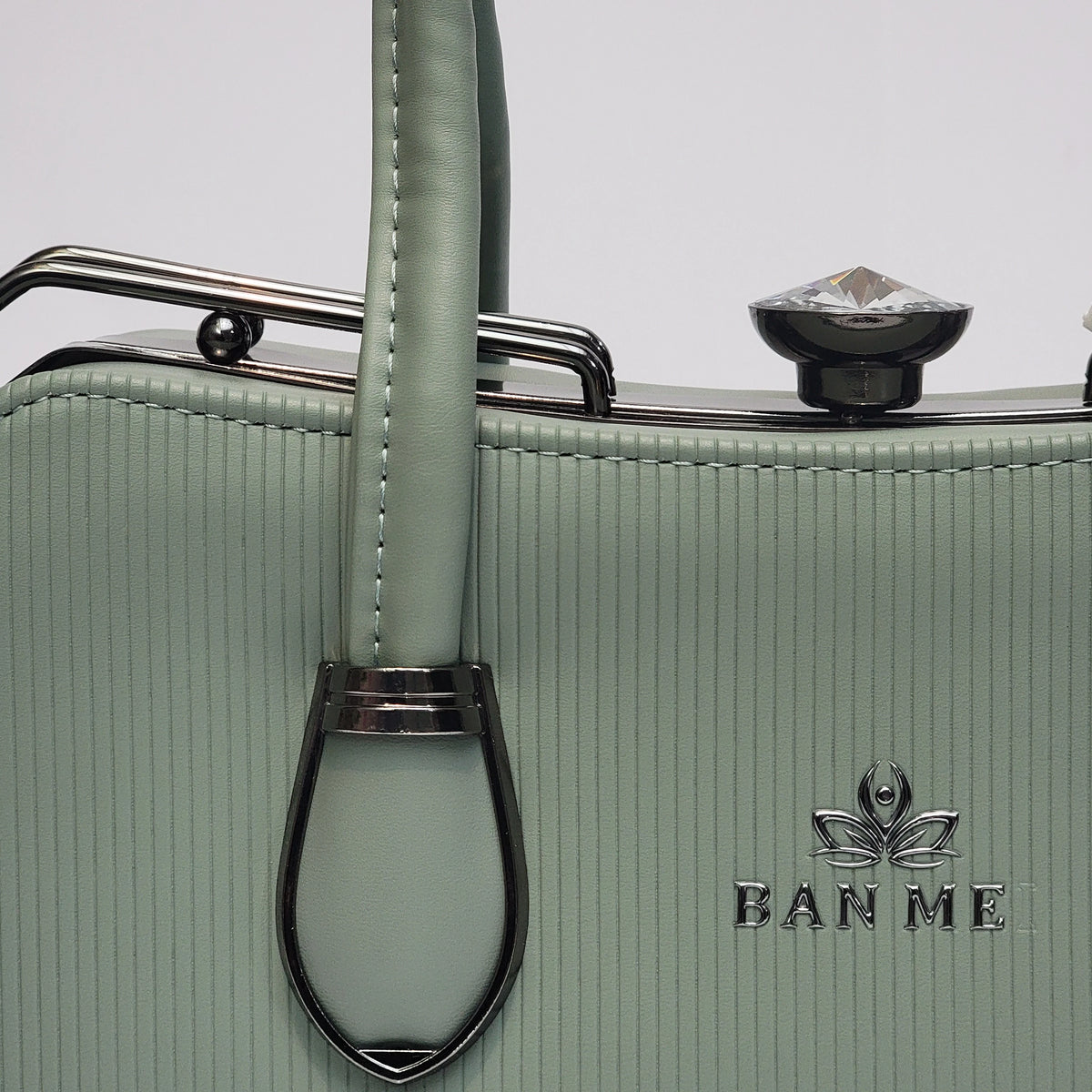 Close-up of a sage green textured handbag with a vertical handle, silver-tone metal frame, and the BAN ME logo on the front.