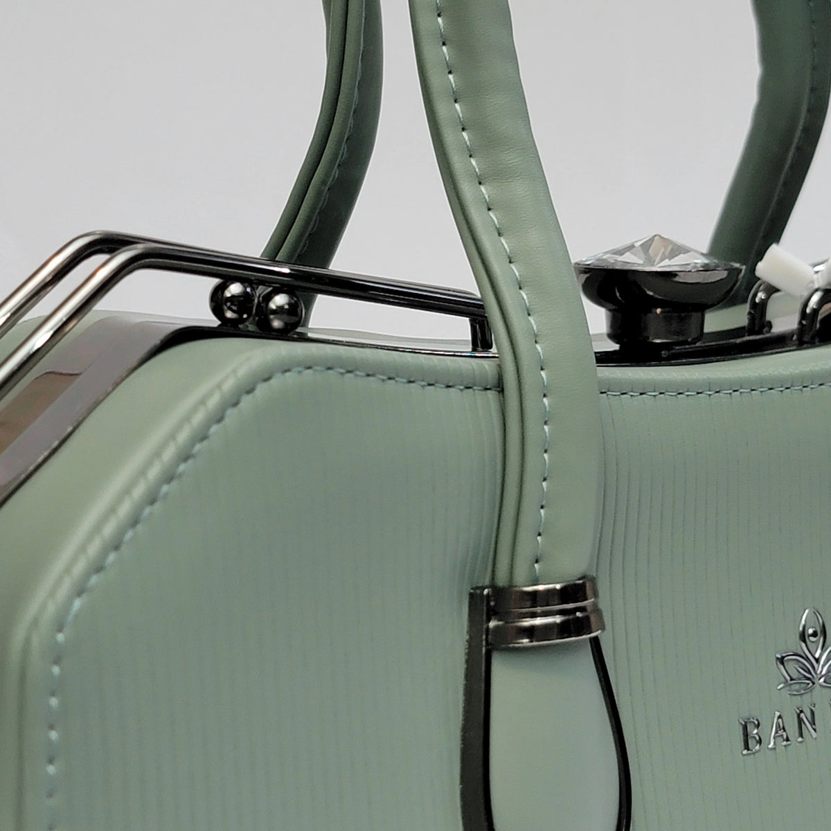 Close-up of a mint green textured leather handbag with silver-tone metal hardware, a decorative crystal accent, and a silver embroidered logo on the side.