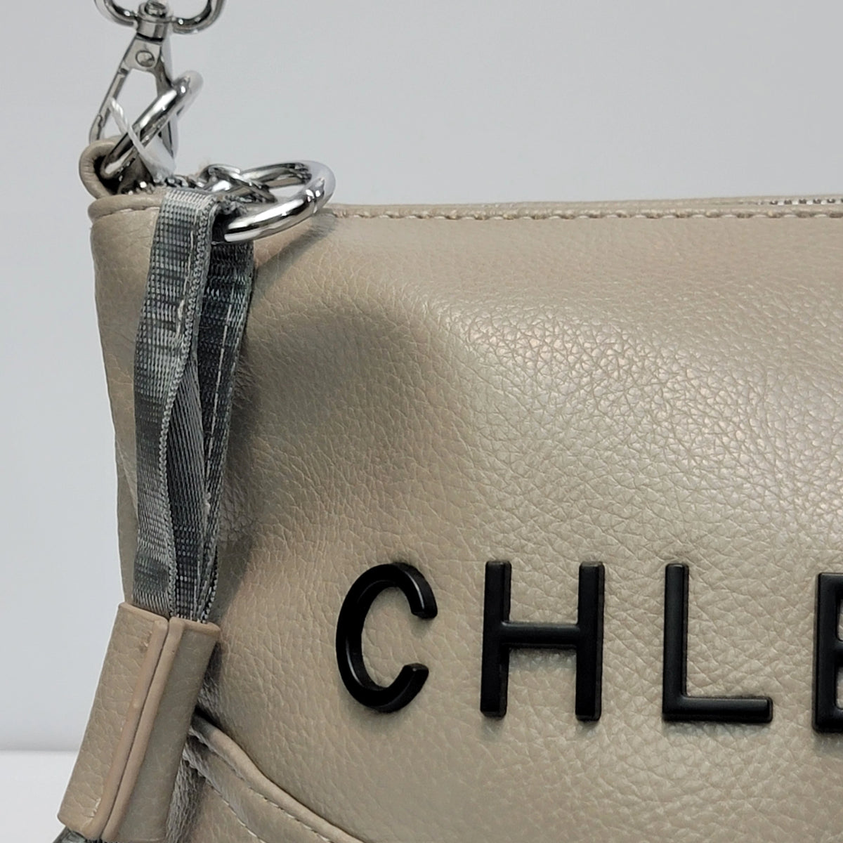 Close-up of a light beige pebbled leather handbag with black CHANEL lettering on the front and a gray woven strap detail with silver hardware.