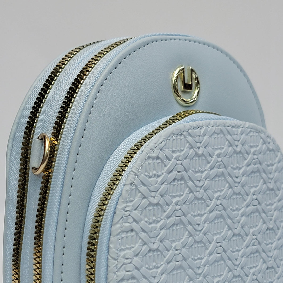 Close-up of a light blue backpack or mini bag with gold-tone zippers and hardware, showing a smooth leather-like top panel and a textured front pocket.