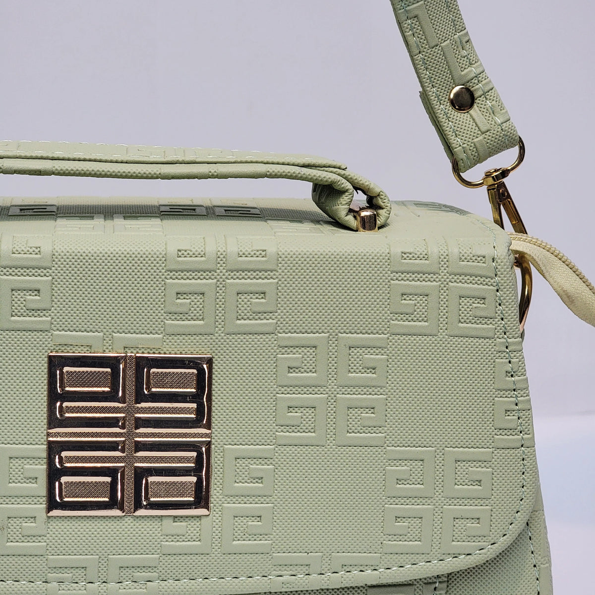 A close-up of a light sage green Givenchy handbag featuring the brand's signature embossed Greek key pattern and a metallic gold-toned four-square logo clasp on the front flap.