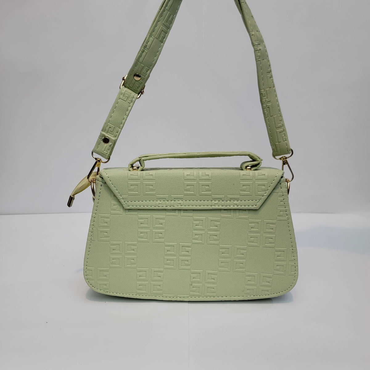A light sage green handbag with an embossed geometric monogram pattern, featuring a top handle, a shoulder strap, and gold-tone hardware, set against a plain light gray background.