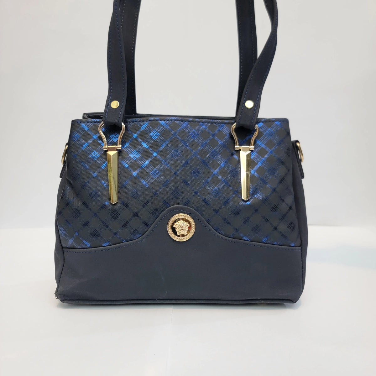 A structured handbag with a dark navy blue base and a blue and black diamond-patterned quilted front flap, featuring gold-tone hardware and a circular gold-tone medallion on the front.