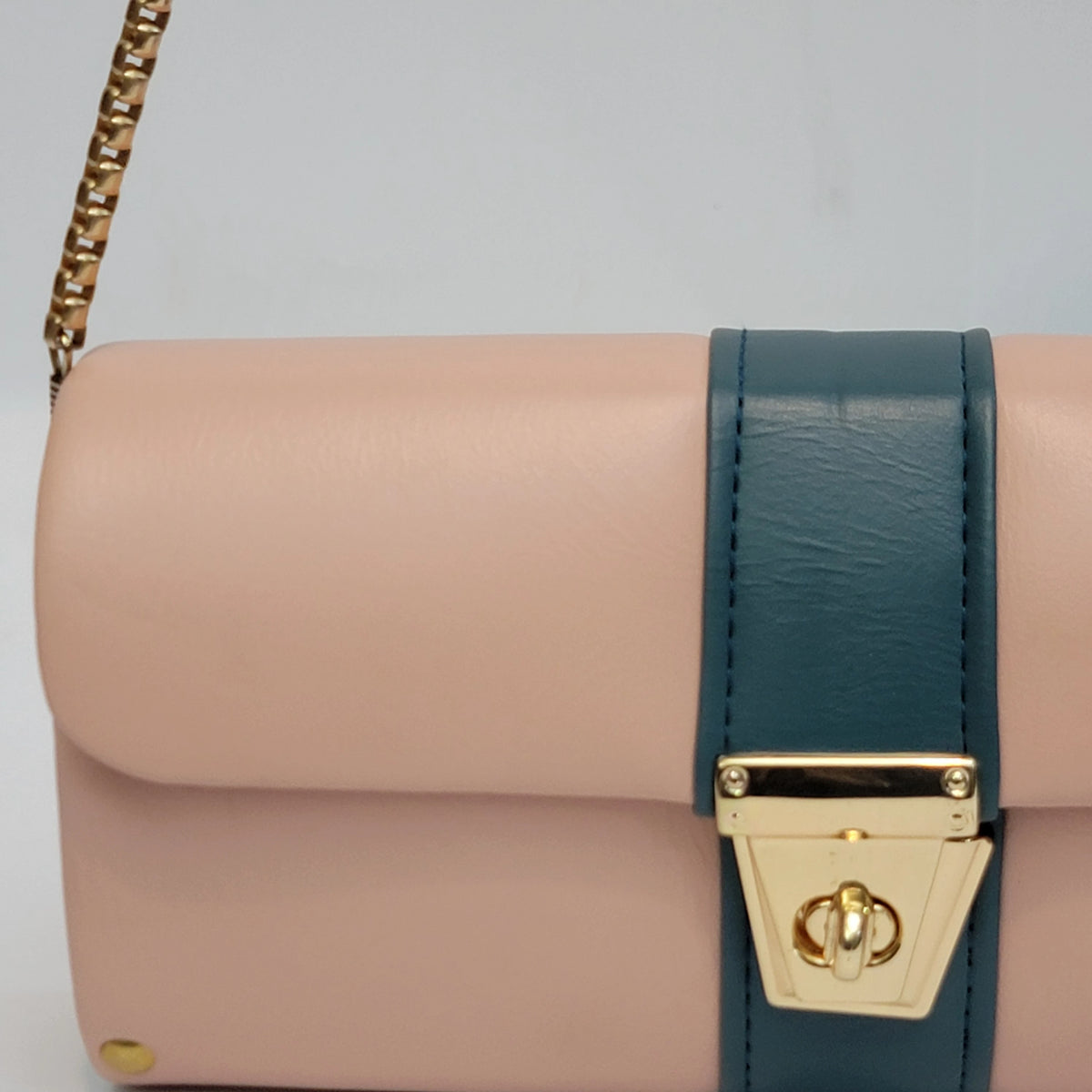 A close-up of a handbag with a light pink leather body, a dark teal vertical strap, and a gold-tone turn-lock clasp and chain strap.