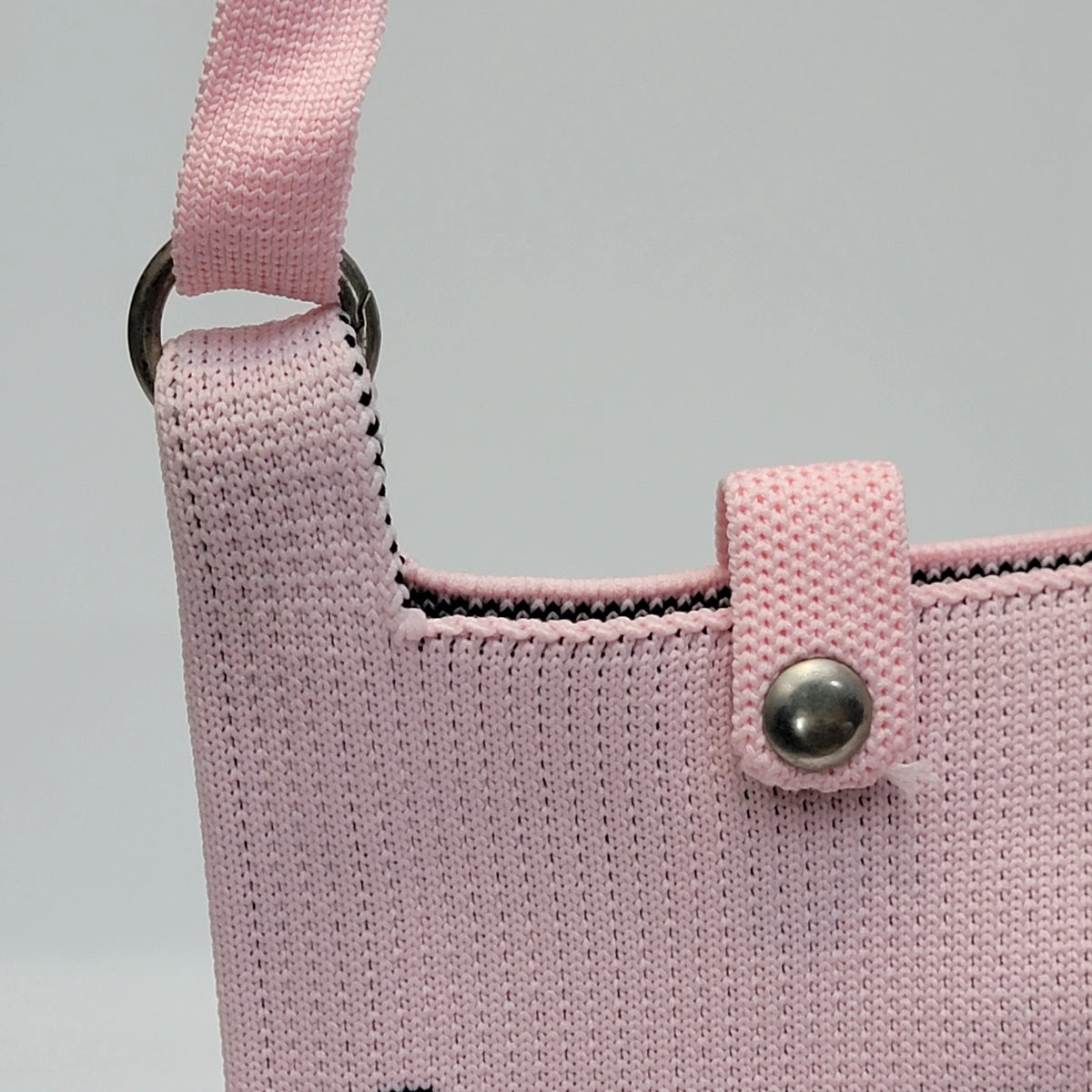 Close-up of a light pink knitted shoulder bag with a matching pink strap, metal ring, and silver snap button closure, set against a neutral gray background.
