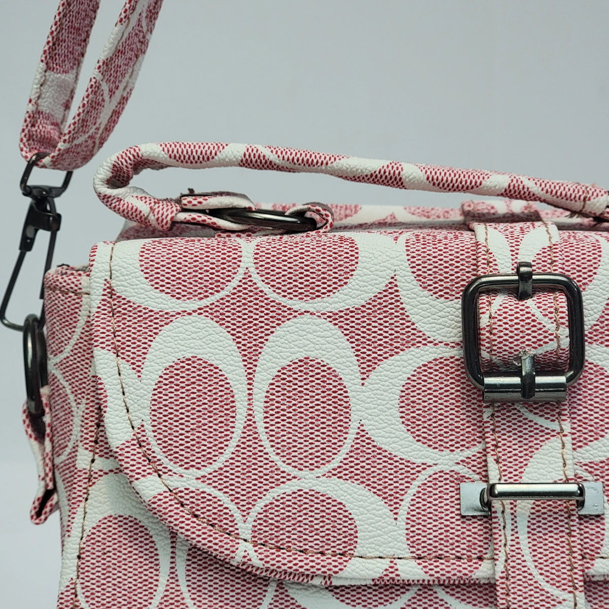 A close-up of a handbag featuring a pink and white monogram pattern, with a top handle, flap closure, and silver-tone hardware against a light gray background.