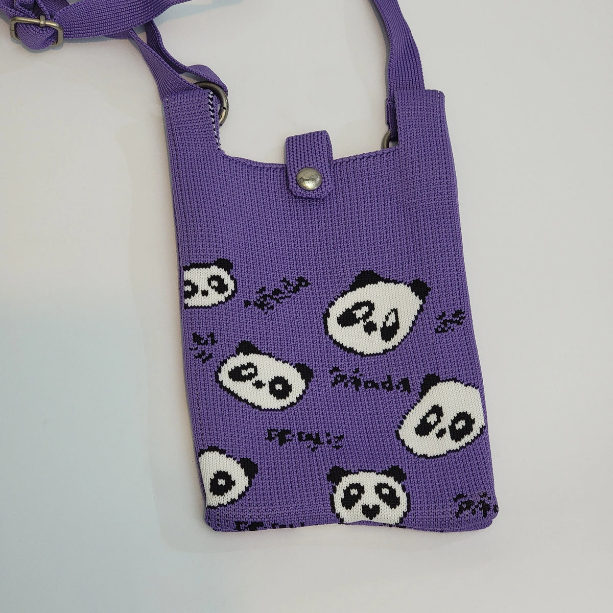 A purple knitted crossbody bag featuring a playful pattern of black and white panda faces and handwritten text, shown on a plain light background.