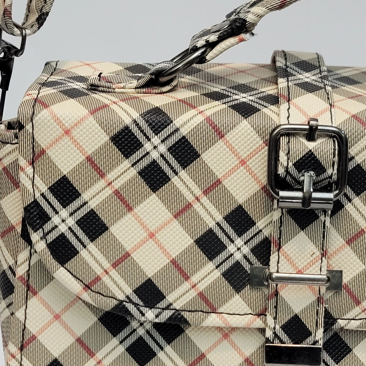 Close-up of a plaid shoulder bag with a beige, black, and white tartan pattern accented by thin red lines. The bag features a front flap with a large silver buckle and a matching woven strap.