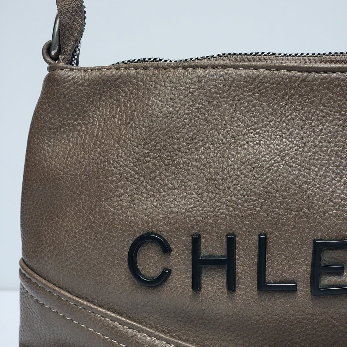 Close-up of a taupe pebbled leather handbag with black CHANEL lettering on the front, a top zipper, and a matching leather strap against a light gray background.