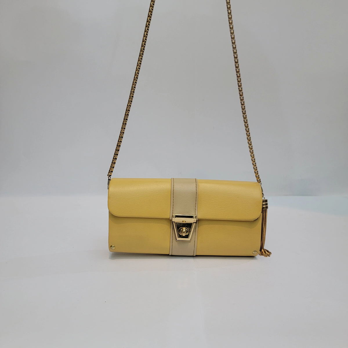 A yellow leather shoulder bag with a beige center strap and gold-tone hardware, including a chain strap and a small tassel detail, shown against a plain light gray background.