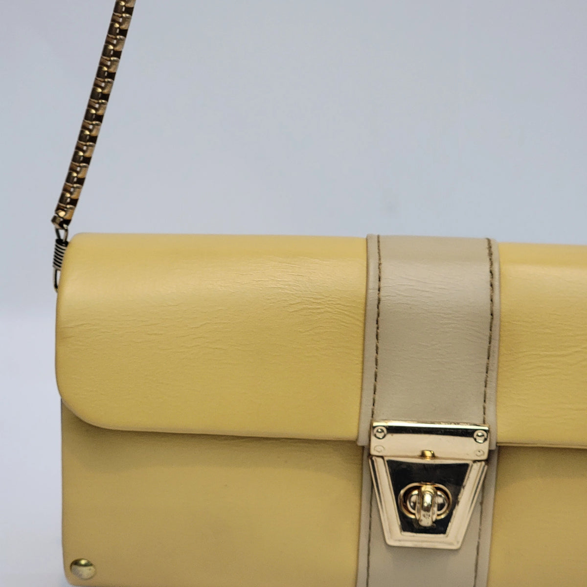 A close-up of a yellow leather shoulder bag with a beige vertical strap and gold-tone hardware, including a turn-lock clasp and a gold chain strap.